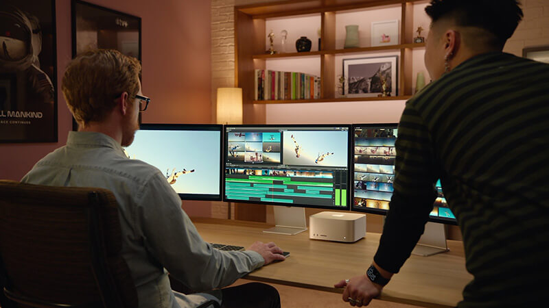mac studio_multiple_displays Deux monteurs examinent une ligne de temps sur trois moniteurs reliés à un studio Mac.