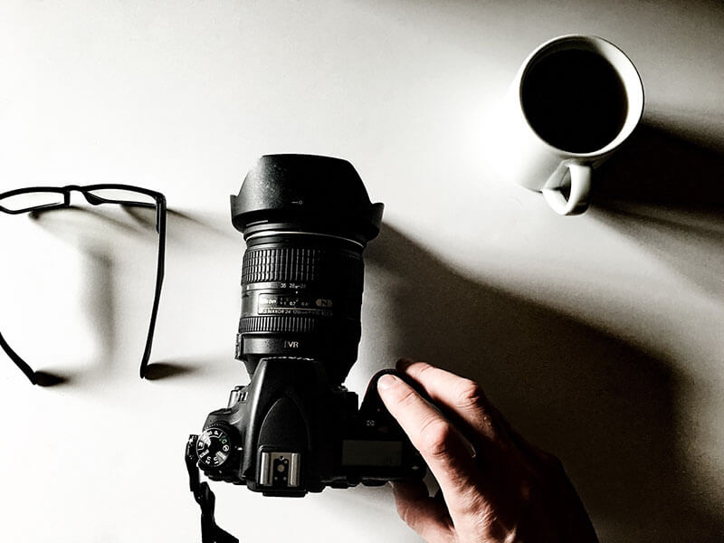 lateral-caracoles-cámara-café Un hombre coloca una cámara DSLR en una mesa junto a una taza de café negro y un par de gafas de lectura